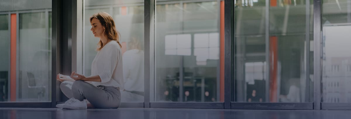 Woman seated in lotus position in an office building with windows.