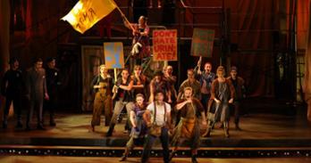 Students perform Urinetown: the Musical, waving protest signs and singing on dark stage in industrial work clothes.