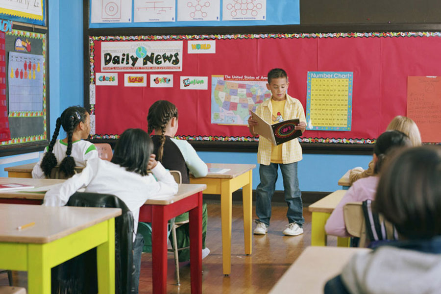 Child stands at front of classroom, reading a book aloud