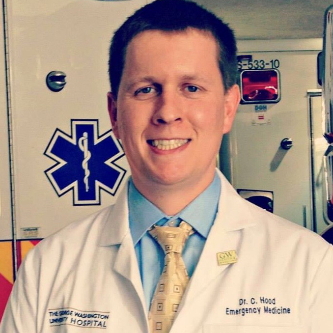 A man is smiling in front of a blue medical sign, wearing a white lab coat, blue shirt, and yellow tie.