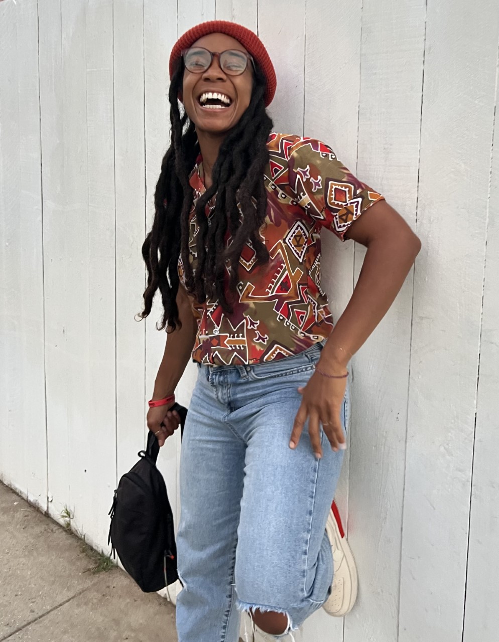 smiling person with long black hair in brown shirt and blue jeans leaning against a white fence.