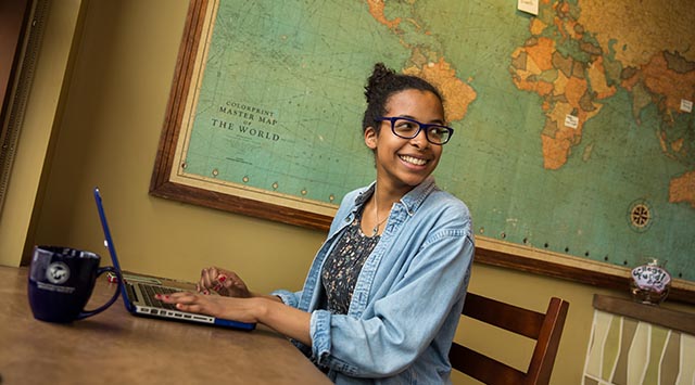 smiling girl sits at a laptop with coffee mug and a map on the wall behind her