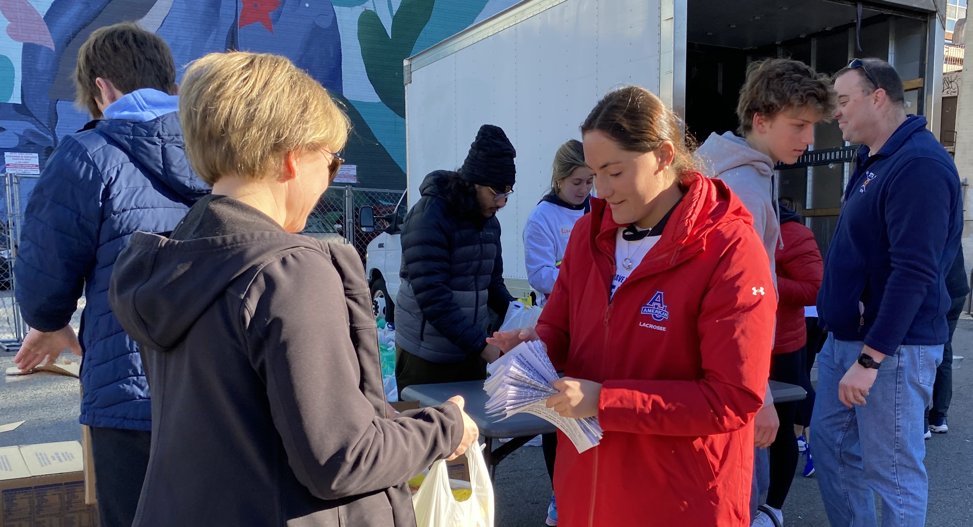 AU student in red coat hands out food to community member.