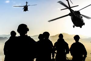 American soldiers underneath two helicopters, looking at mountains in the distance.